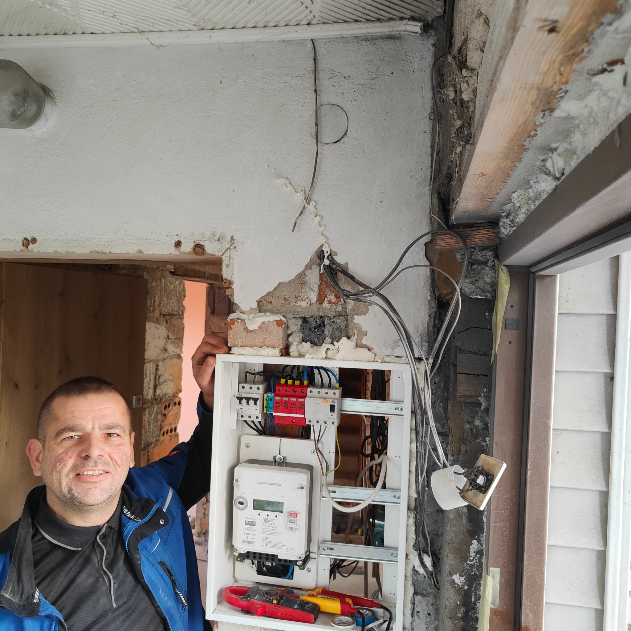 Elektryk z Łuczyny prezentuje zamontowaną skrzynkę elektryczną z licznikiem i zabezpieczeniami w trakcie prac wykończeniowych, widoczne przewody, ściany w stanie surowym i narzędzia pomiarowe.