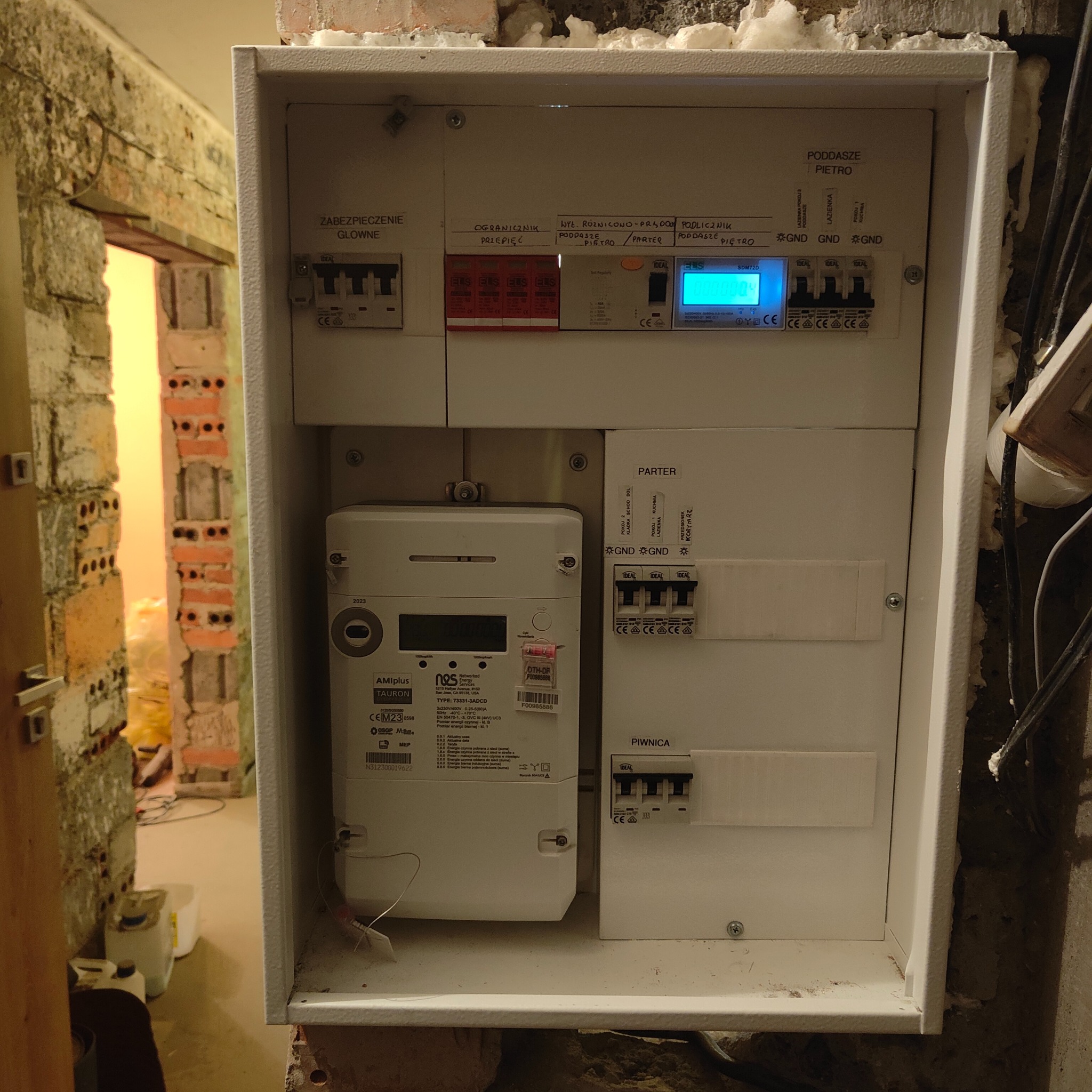 Rozdzielnica elektryczna z zamontowanym licznikiem energii elektrycznej w trakcie remontu, widoczne przewody i surowa ściana z cegły