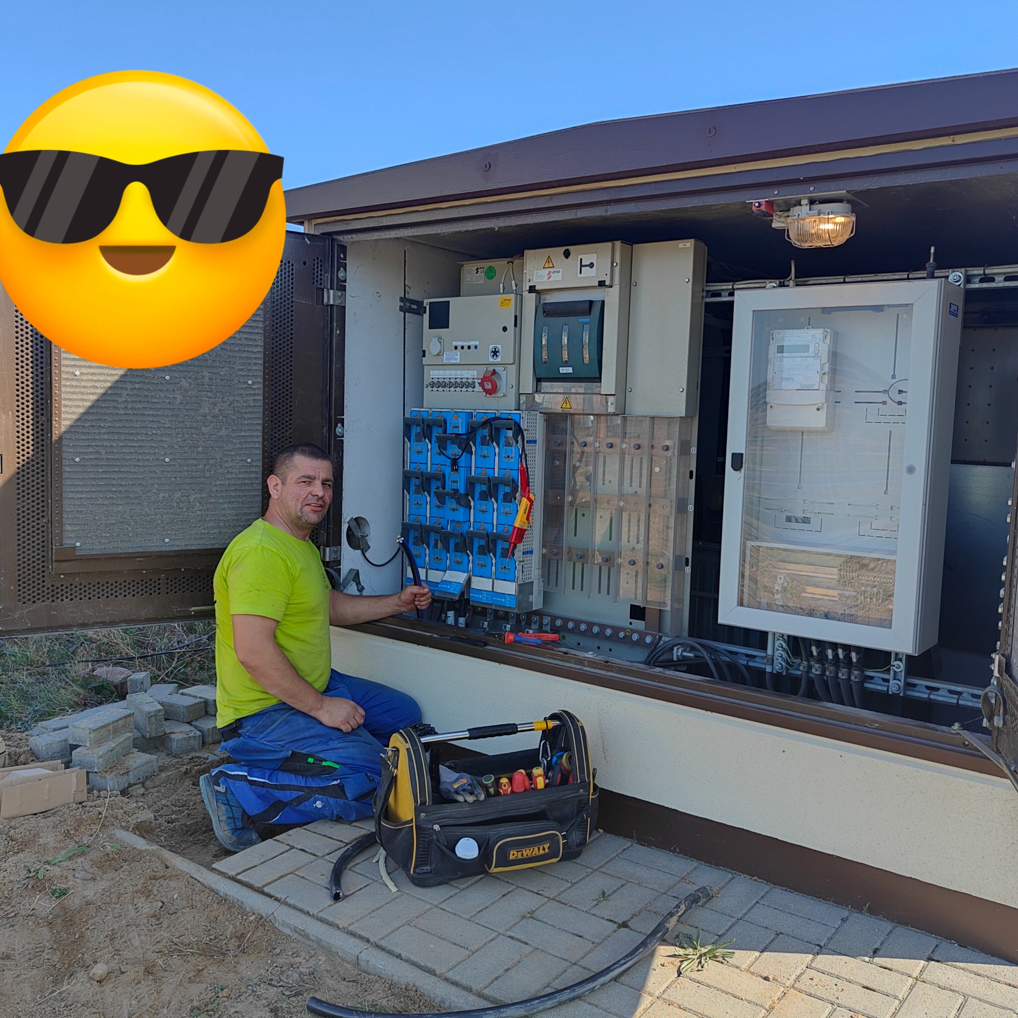 Elektryk w zielonej koszulce, klęczący przed otwartą szafą elektryczną z bezpiecznikami i licznikiem, trzyma kombinerki, obok torba narzędziowa DeWalt.