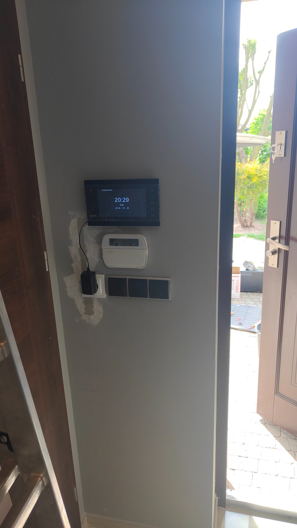 Zamontowany panel sterowania domofonem z widocznym datą i godziną, obok centrala alarmowa i włączniki światła na szarej ścianie, widoczne ślady po montażu, otwarte drzwi wejściowe z widokiem na ogród.