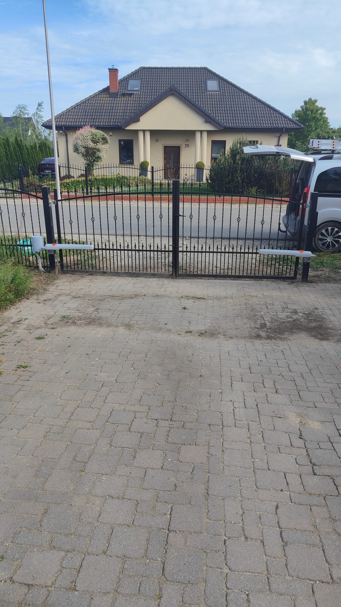 Czarna, metalowa brama wjazdowa z automatyką, widok na dom z beżową elewacją i szarym dachem, podjazd z kostki brukowej.