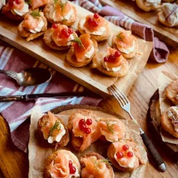 Mini kanapeczki z łososiem, kremowym serkiem i czerwoną porzeczką, podane na drewnianej desce z dekoracyjnymi sztućcami i ściereczką w paski.