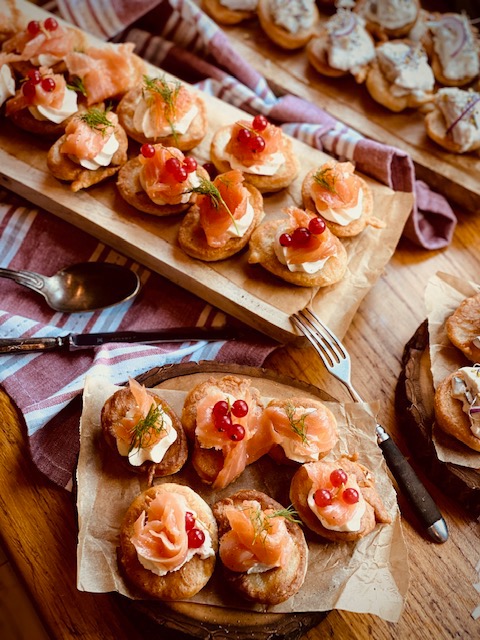 Mini kanapeczki z łososiem, kremowym serkiem i czerwoną porzeczką, podane na drewnianej desce z dekoracyjnymi sztućcami i ściereczką w paski.