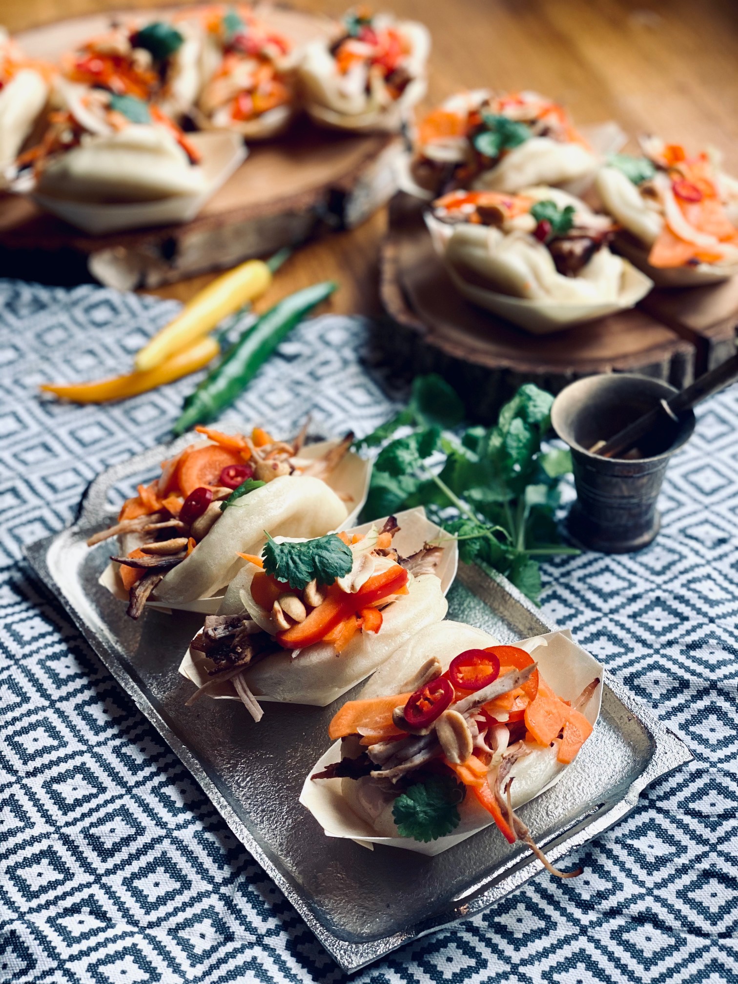 Elegancko podane bao buns z warzywami i orzechami na srebrnej tacy, rustykalne drewniane podstawki w tle, całość na wzorzystej tkaninie.