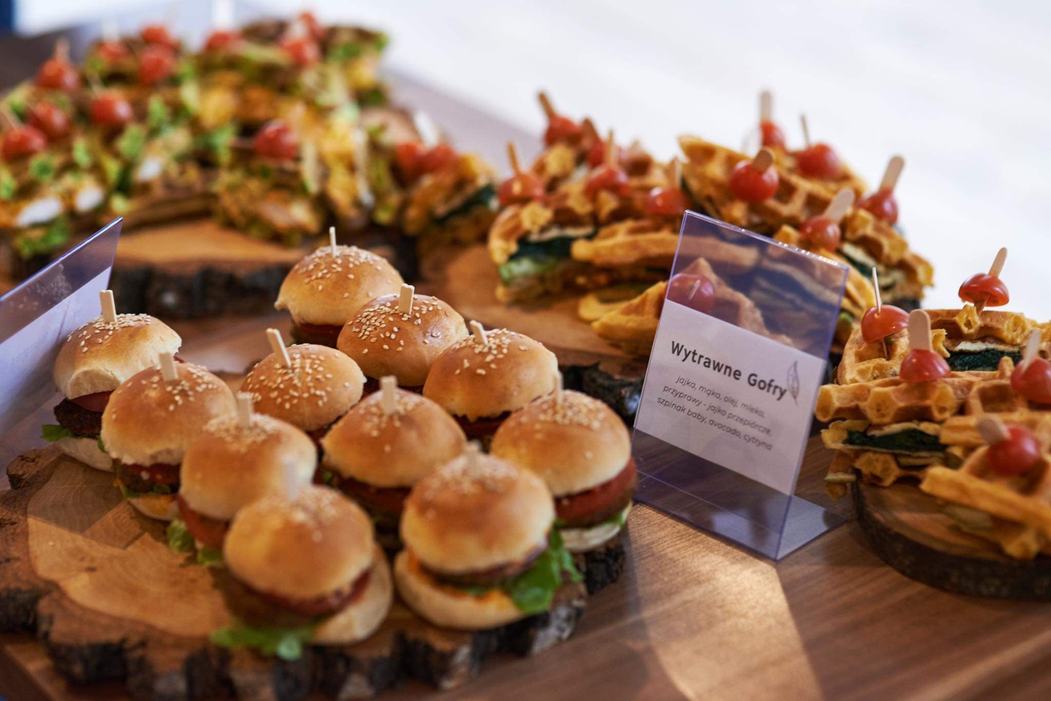 Mini burgery z sezamem i wytrawne gofry z pomidorkami koktajlowymi na drewnianych podstawkach, prezentacja menu na przyjęciu.