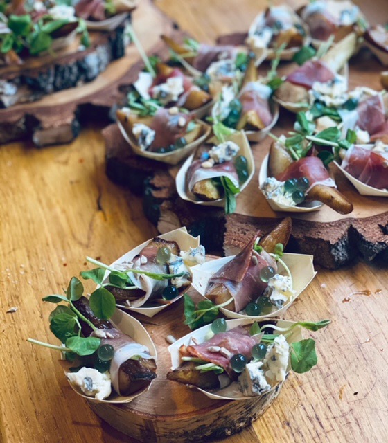 Eleganckie przekąski cateringowe: gruszka owinięta szynką parmeńską, z serem pleśniowym, orzeszkami piniowymi i zielonymi koralikami na drewnianych łódeczkach, ułożone na rustykalnej desce z kory.