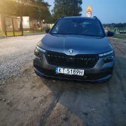 Szary samochód osobowy marki Skoda Kamiq z polską tablicą rejestracyjną KT 5189H, zaparkowany na piaszczystym terenie z widocznymi śladami opon, w tle znak drogowy ostrzegawczy i zabudowania.