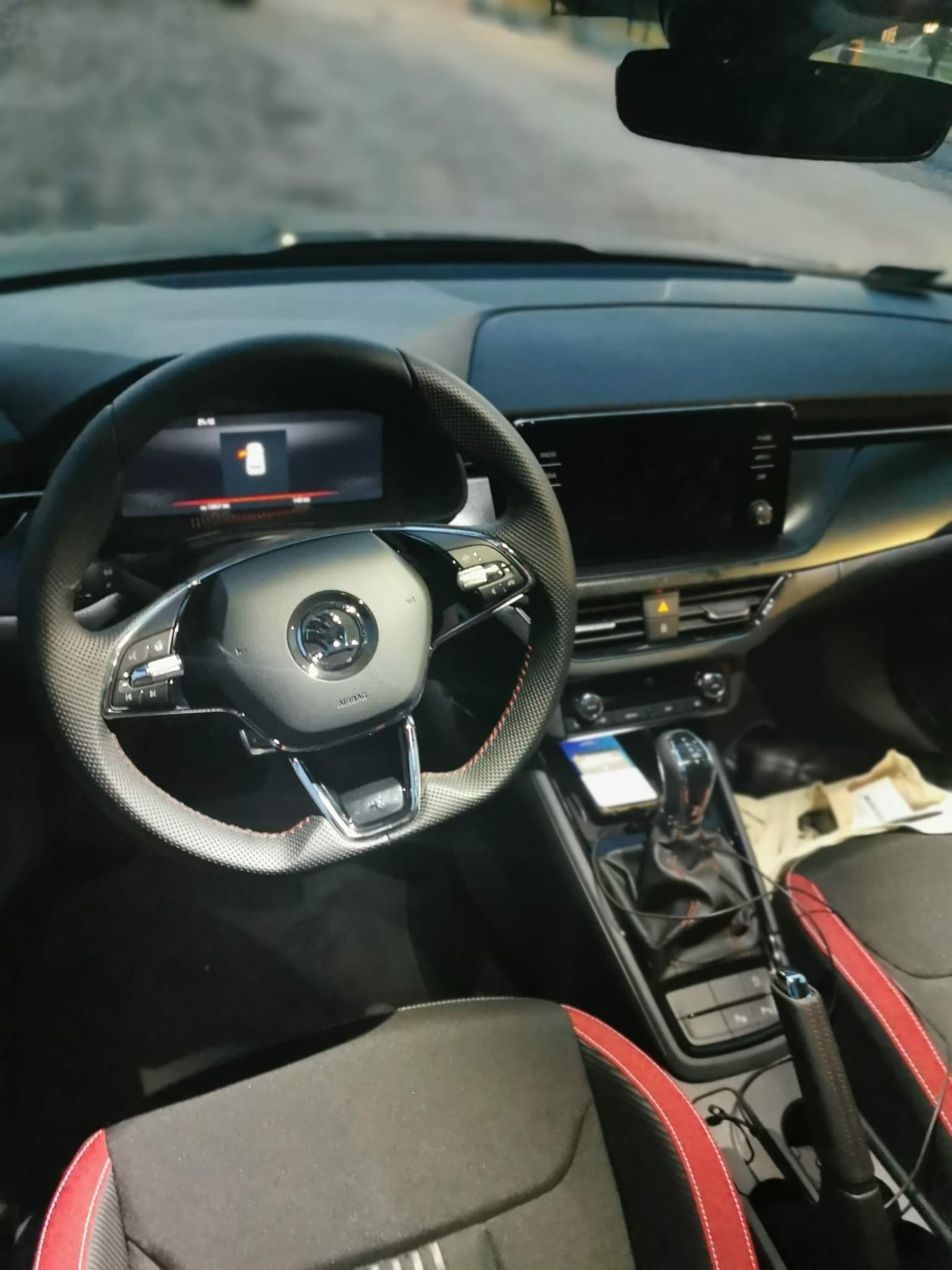 Wnętrze samochodu marki Skoda z czarną tapicerką i czerwonymi akcentami, widoczna kierownica z logo i deska rozdzielcza z ekranami.