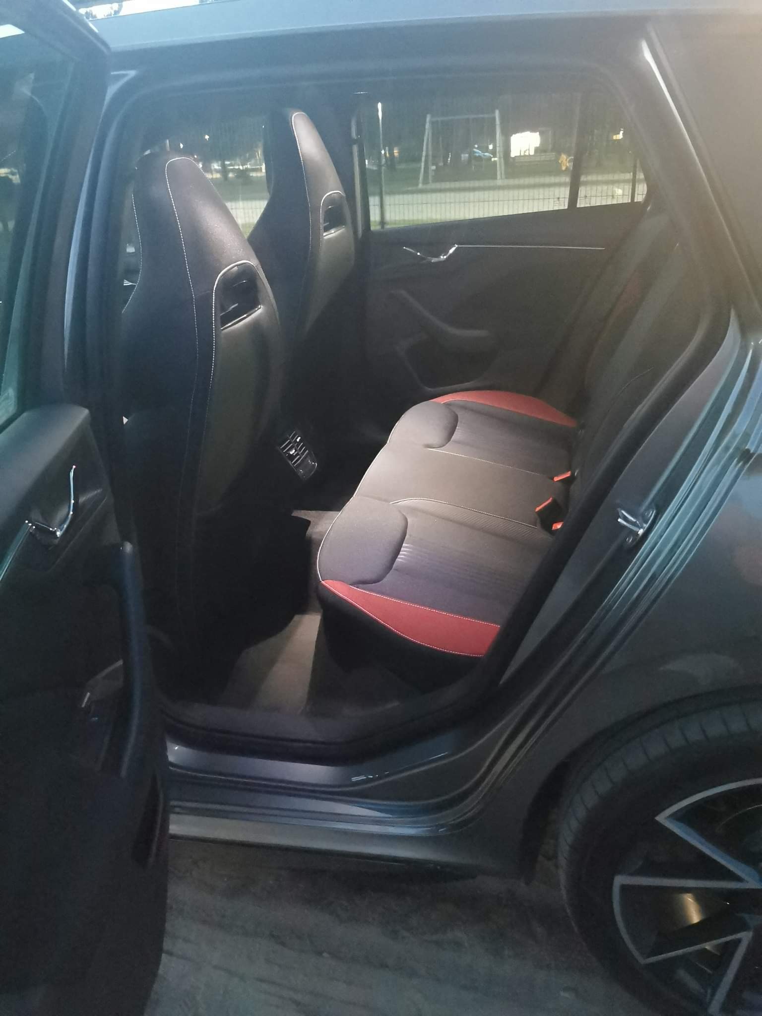 Wnętrze samochodu z otwartymi drzwiami, widoczne czarne sportowe fotele z białymi przeszyciami i tylna kanapa z czerwonymi akcentami.