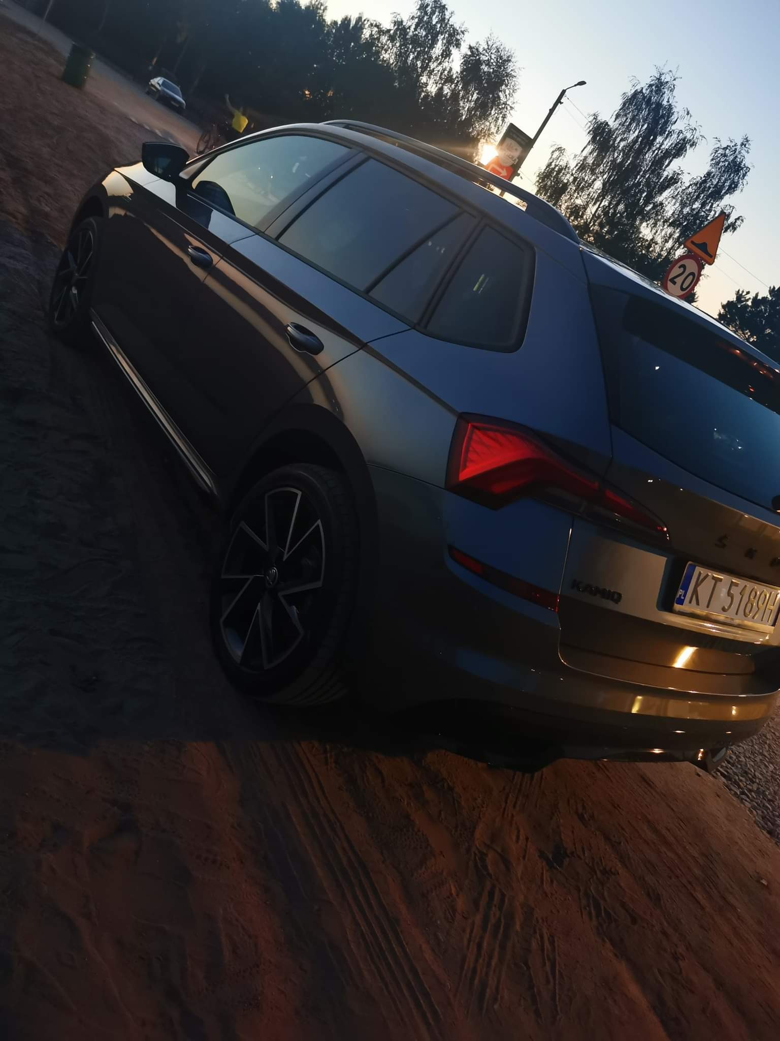 Szary samochód kombi marki Skoda Kamiq zaparkowany na piaszczystym terenie, widok z tyłu, z widoczną tablicą rejestracyjną KT 5189H oraz znakiem ograniczenia prędkości do 20 km/h w tle.