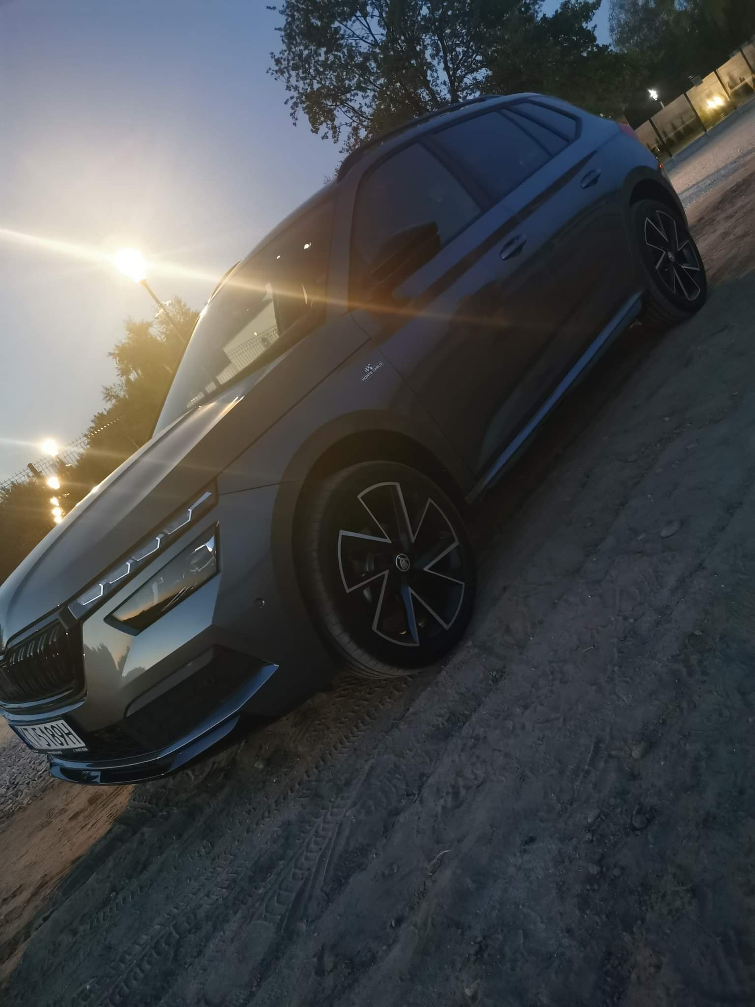 Szary samochód Skoda Kamiq zaparkowany na piaszczystym podłożu, widok z boku, oświetlony wieczornym słońcem, z widocznymi detalami felg i przednich świateł.