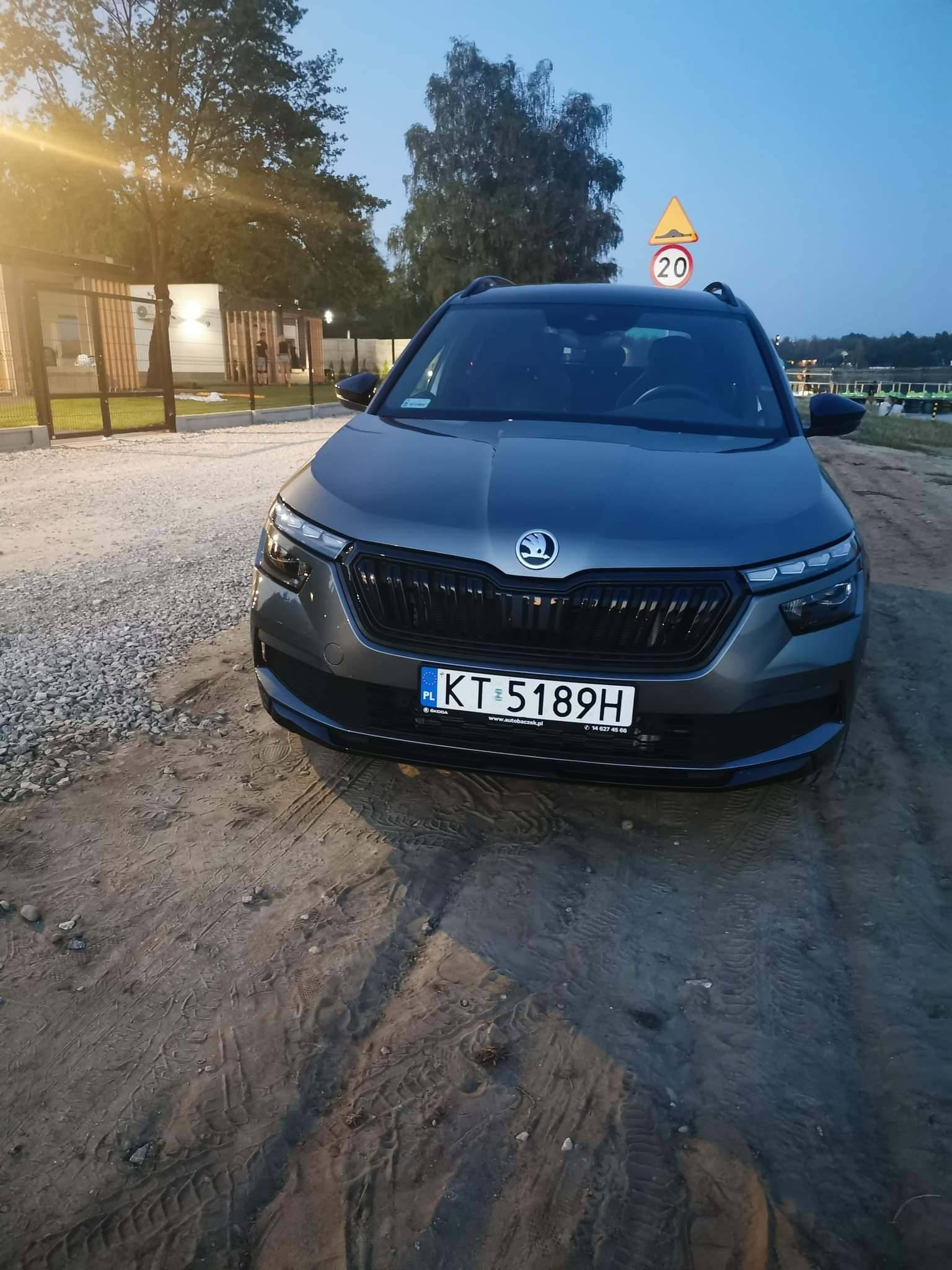 Szary samochód osobowy marki Skoda Kamiq z polską tablicą rejestracyjną KT 5189H, zaparkowany na piaszczystym terenie z widocznymi śladami opon, w tle znak drogowy ostrzegawczy i zabudowania.