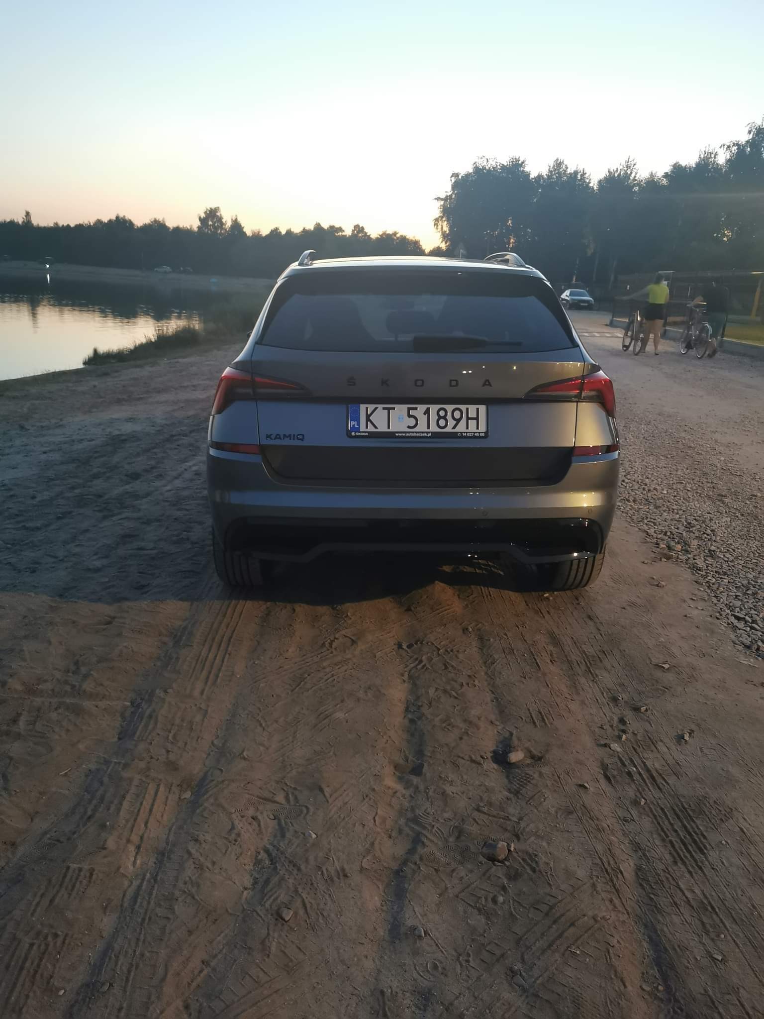 Szary samochód Skoda Kamiq z polskimi tablicami rejestracyjnymi KT 5189H, zaparkowany tyłem na piaszczystej drodze obok jeziora o zmierzchu.