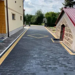 Ułożona, ciemnoszara kostka brukowa z żółtymi akcentami tworząca wzór rombu na podjeździe przed budynkiem z kremową elewacją i małym budynkiem z kamienia i czerwoną blachodachówką.