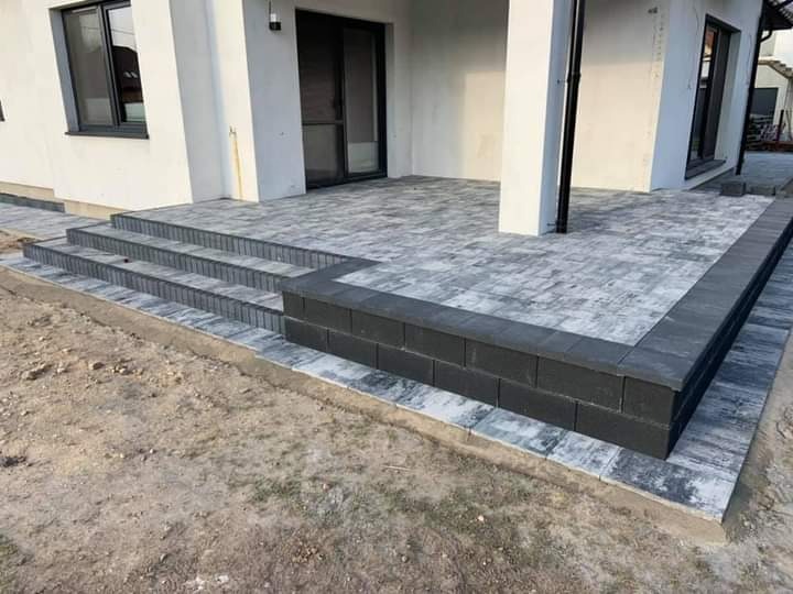 Nowoczesny taras z kostki brukowej w odcieniach szarości, z trzema schodami prowadzącymi do domu o białej elewacji.