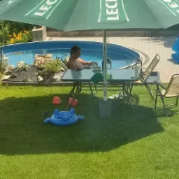 Osoba relaksuje się przy basenie w ogrodzie, siedząc pod parasolem obok stołu z krzesłami, w tle widoczne rośliny i zabawki dla dzieci na trawniku.