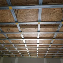 Sufit podwieszany z metalową konstrukcją nośną, przymocowaną do drewnianej płyty OSB, widoczne żółte taśmy mocujące i fragment drabiny malarskiej.