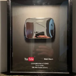 Oprawiona srebrna nagroda YouTube za przekroczenie 100 000 subskrybentów, z widocznym logo YouTube i napisem 'Congratulations', odbicie osoby robiącej zdjęcie w błyszczącej powierzchni nagrody.