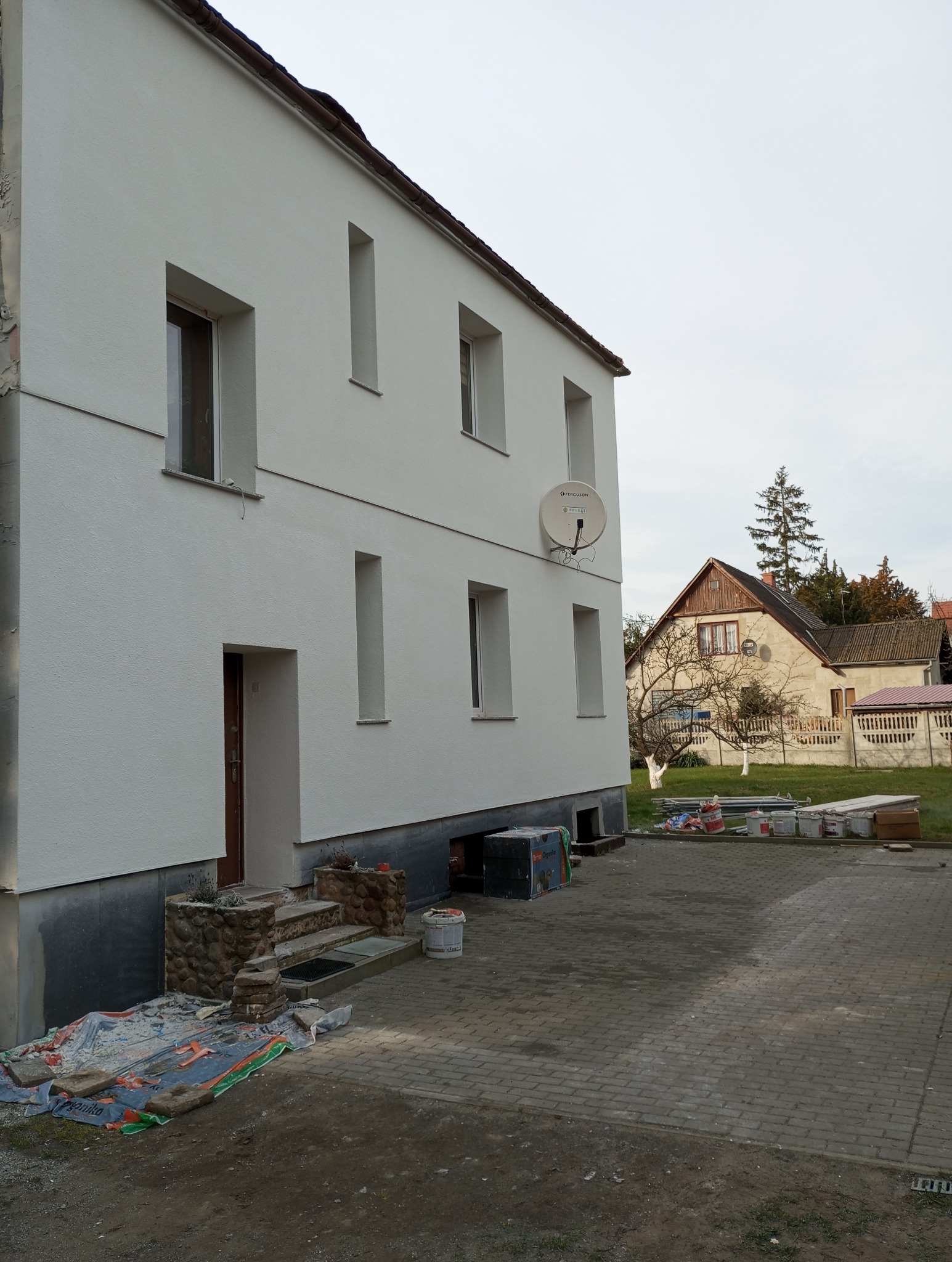 Biała elewacja dwukondygnacyjnego domu z brązowymi oknami i satelitą. Widoczne materiały budowlane i narzędzia. W tle drugi dom i drzewa.