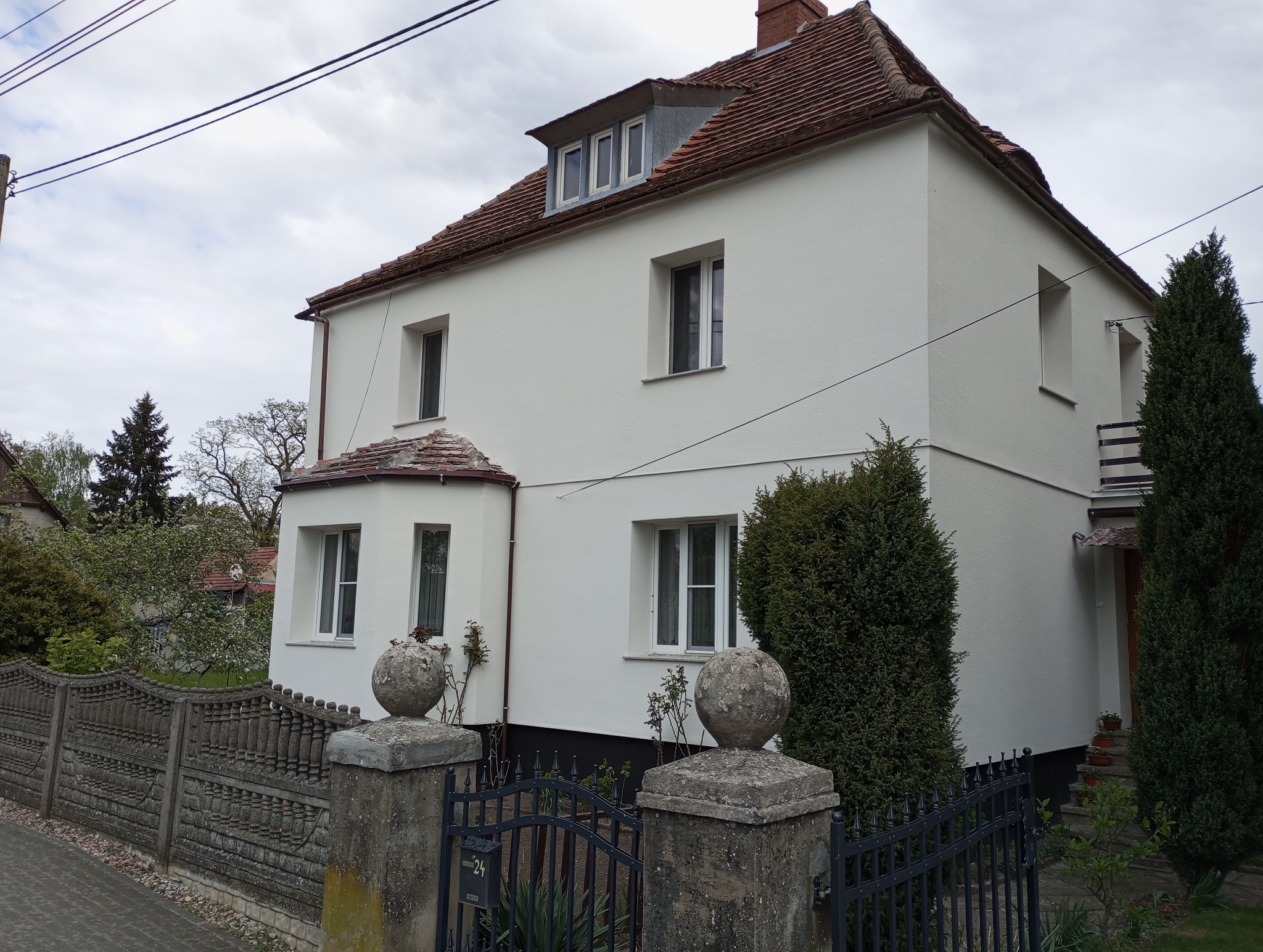 Biały dom z czerwoną dachówką, widoczny zza ozdobnego, metalowego ogrodzenia. Elewacja po renowacji, z widocznymi detalami architektonicznymi i zadbanym otoczeniem.