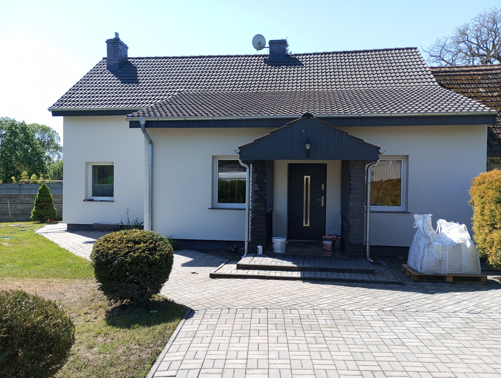 Dom jednorodzinny z jasną elewacją i ciemnym dachem. Na podjeździe worki z materiałami budowlanymi. Widoczne świeżo pomalowane ściany i odnowione wejście.
