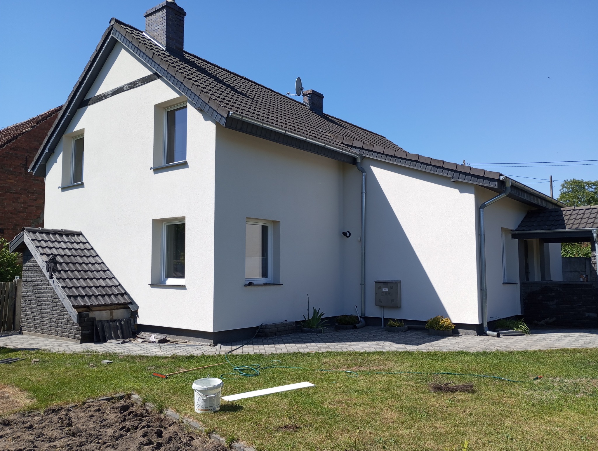 Biała elewacja domu jednorodzinnego w słoneczny dzień. Widoczne okna, dach z dachówki, trawnik i elementy ogrodu. Świeżo pomalowana fasada.