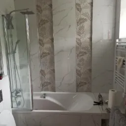 Wykończona łazienka z wanną obudowaną marmurowymi płytkami, kabiną prysznicową ze szklanymi drzwiami, grzejnikiem drabinkowym i dekoracyjnymi panelami z motywem liści, widoczny silikon i ręcznik.