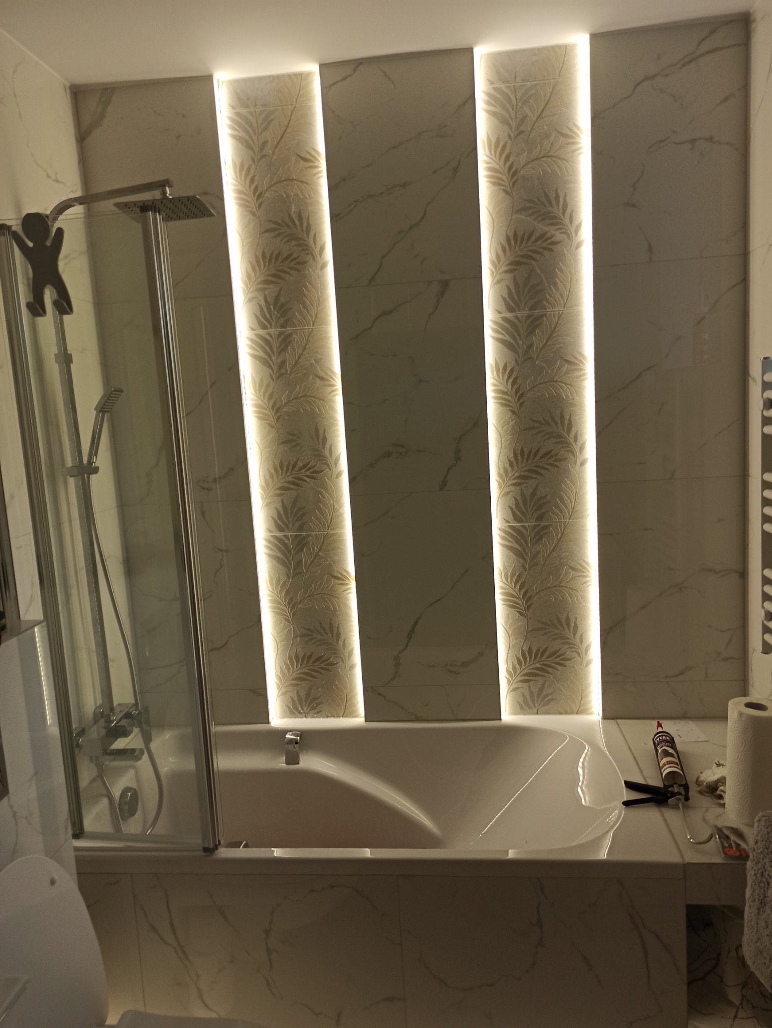 Wykończona łazienka z wanną obudowaną płytkami imitującymi marmur, podświetlane pionowe pasy płytek z motywem roślinnym, kabina prysznicowa ze szkła, na obudowie wanny leżą narzędzia i akcesoria...