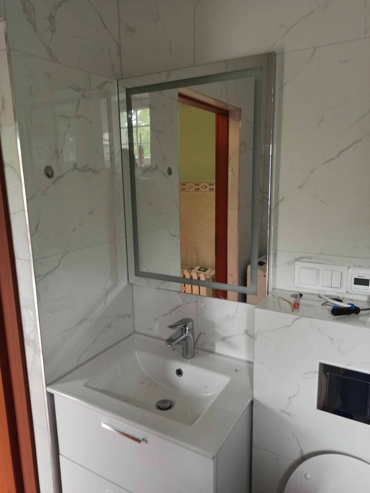 Wykończona łazienka w Rzeszowie z umywalką w szafce, podświetlanym lustrem i płytkami imitującymi marmur na ścianach. Widoczne narzędzia na blacie obok toalety.