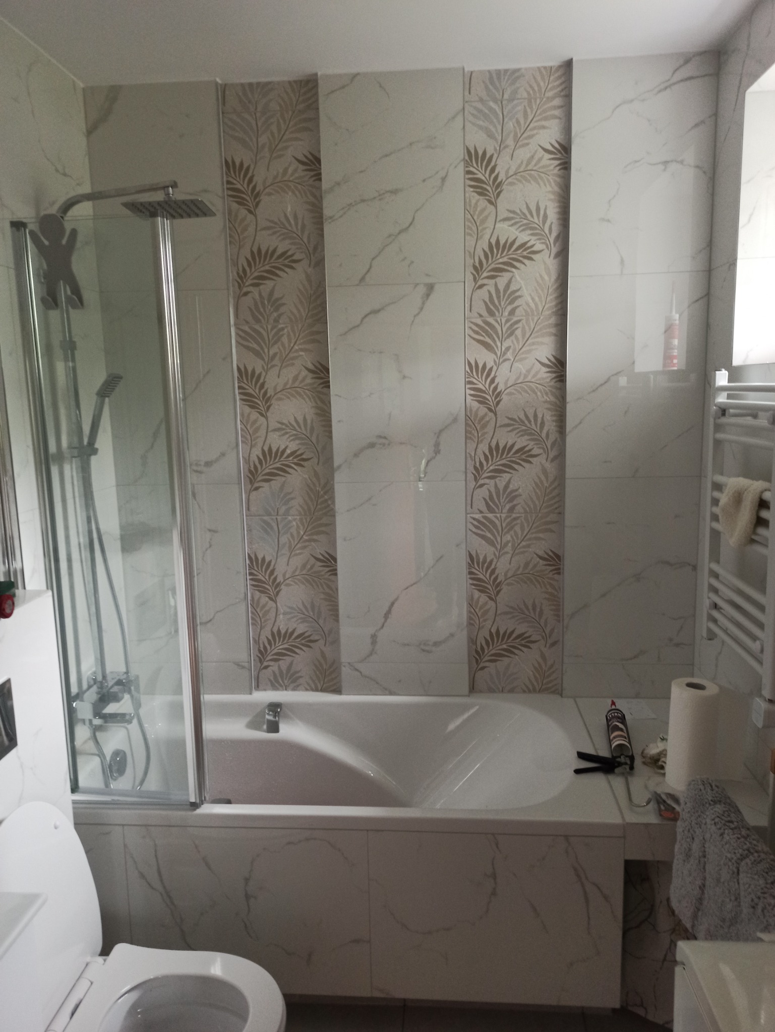 Wykończona łazienka z wanną obudowaną marmurowymi płytkami, kabiną prysznicową ze szklanymi drzwiami, grzejnikiem drabinkowym i dekoracyjnymi panelami z motywem liści, widoczny silikon i ręcznik.