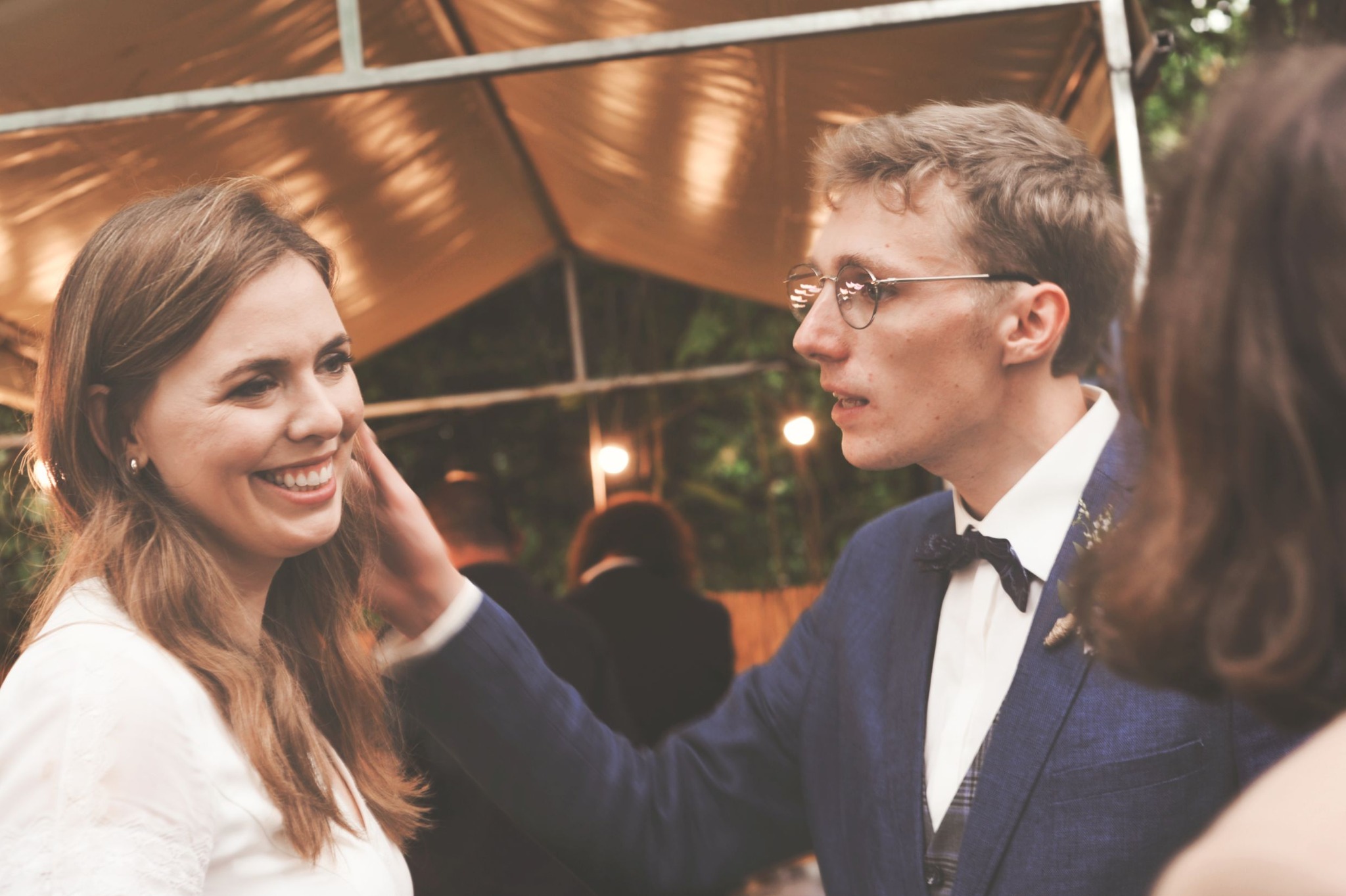 Uśmiechnięta panna młoda z długimi włosami, dotykana przez pana młodego w okularach i granatowym garniturze z muchą, w tle oświetlony namiot.