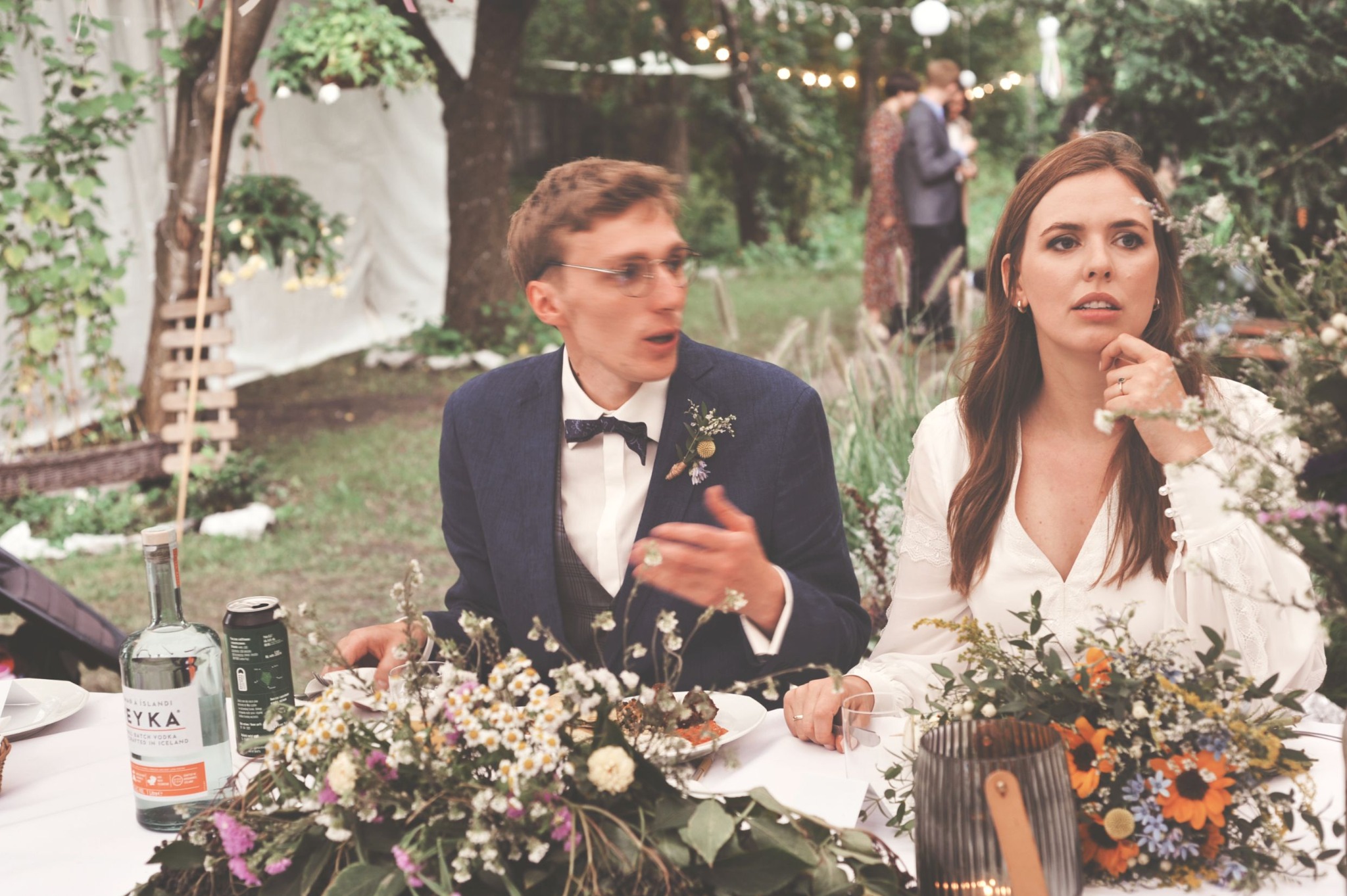 Para młoda siedzi przy stole udekorowanym kwiatami podczas przyjęcia weselnego na świeżym powietrzu, w tle widoczni goście.