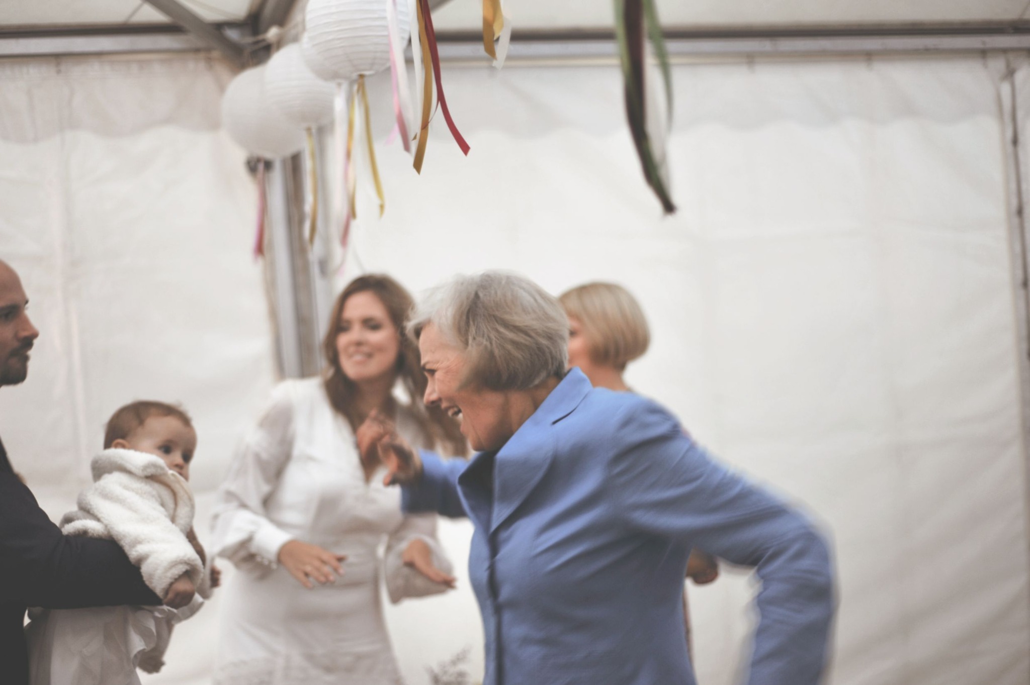 Radosna scena z przyjęcia weselnego: uśmiechnięta kobieta w niebieskiej marynarce tańczy, w tle goście, w tym mężczyzna trzymający dziecko w białym ubranku, całość w jasnym namiocie z dekoracjami...