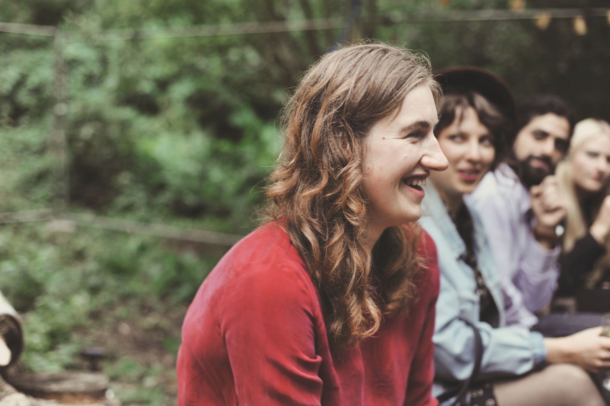 Uśmiechnięta kobieta w czerwonej bluzce z kręconymi włosami, profil, w tle grupa osób na tle zieleni, prawdopodobnie podczas przyjęcia weselnego.