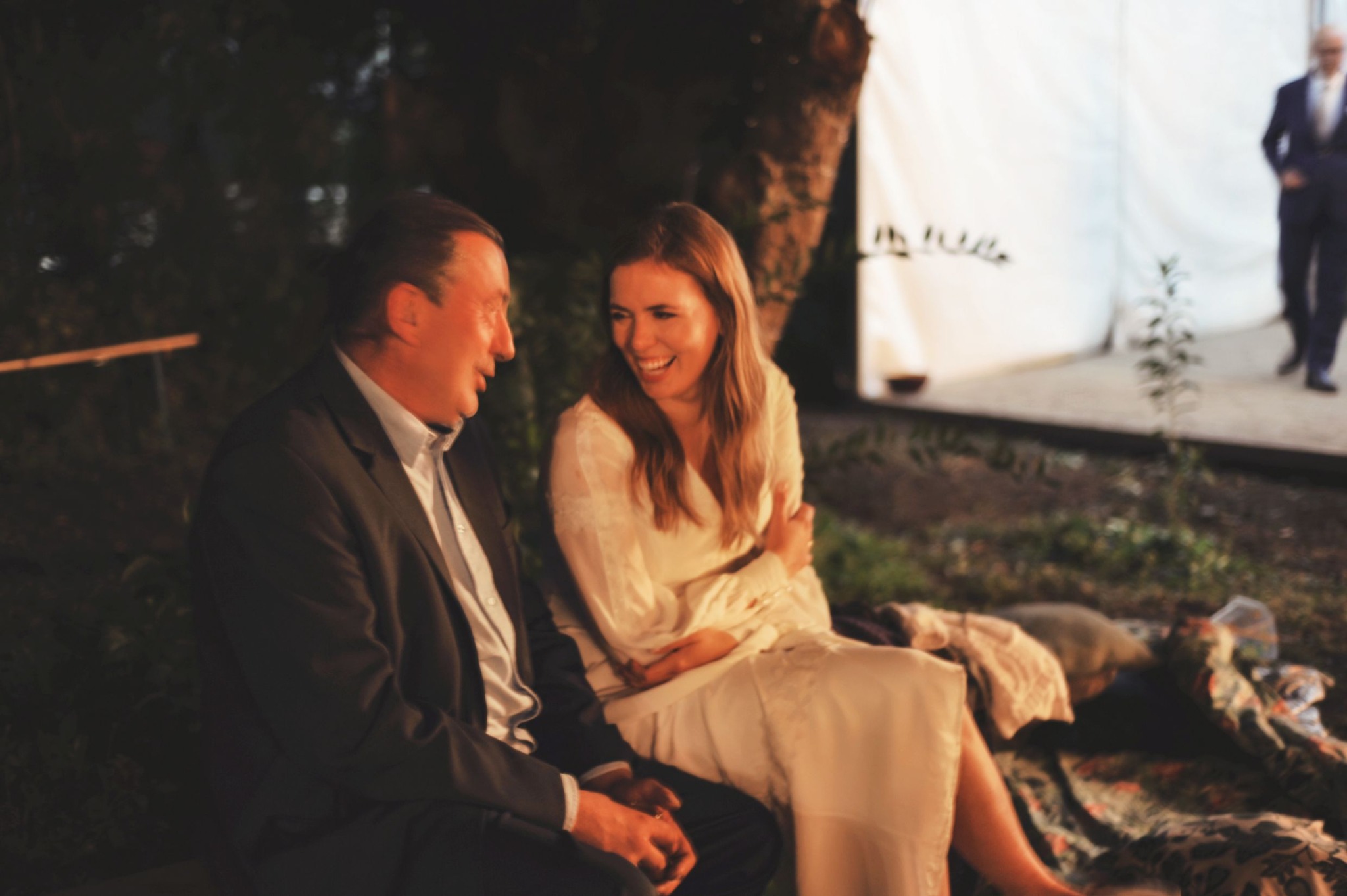 Panna młoda w białej sukni śmieje się rozmawiając z mężczyzną w garniturze podczas reportażu ślubnego, w tle biały namiot i osoba w oddali.