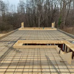 Zbrojenie stropu na drewnianym deskowaniu, widok z góry na siatkę z prętów stalowych, drewniane szalunki i fragment lasu w tle.