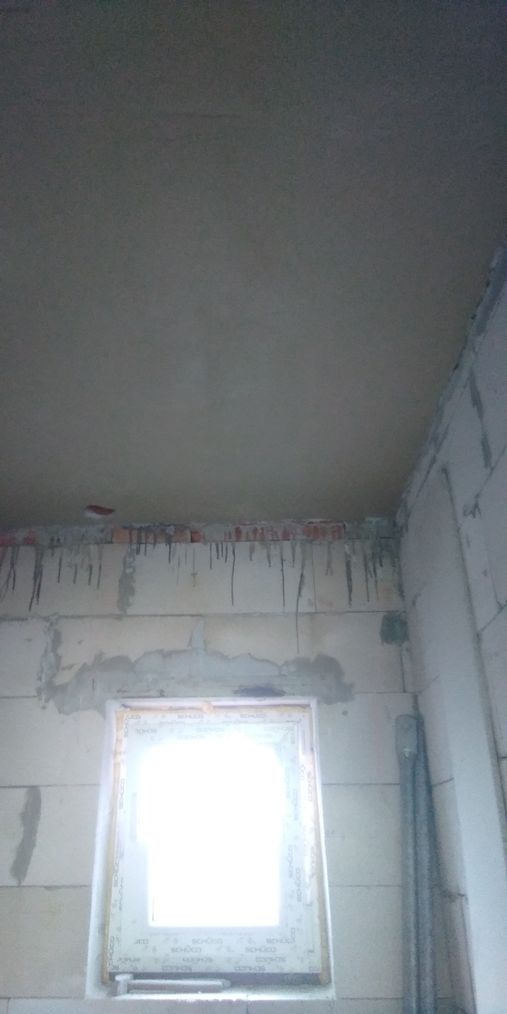 Surowy stan wnętrza: ściany z betonu komórkowego, świeżo położona warstwa tynku na suficie z widocznymi zaciekami, zamontowane okno z profilem PCV, brak wykończenia.