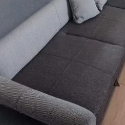 Szary narożnik z poduszkami, widoczne przeszycia i metalowe nóżki, w tle krzesełko do karmienia dziecka.