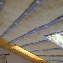 Skośny sufit poddasza z widoczną izolacją z wełny mineralnej, zamocowaną między metalowymi profilami, oraz żółtą folią paroizolacyjną, z oknem dachowym w tle.