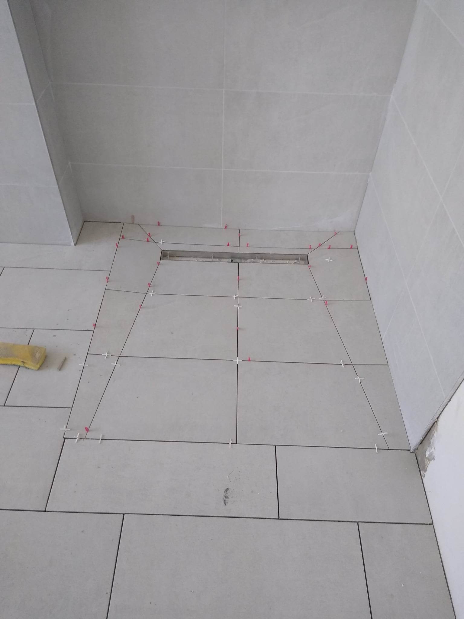 Jasne, prostokątne płytki układane na podłodze i ścianach łazienki, z widocznym odpływem liniowym i krzyżykami dystansowymi w spoinach.