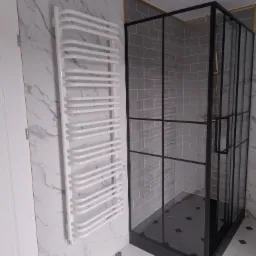 Nowoczesna kabina prysznicowa z czarnymi profilami, szare płytki na ścianach i podłodze, biały grzejnik drabinkowy na ścianie z marmurowym wzorem, biała podłoga z czarnymi akcentami.