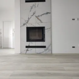 Wykończony kominek obłożony białymi płytkami imitującymi marmur z czarnym wkładem kominkowym i poziomymi wnękami, na podłodze jasnoszara imitacja drewna, widoczne puste gniazdka elektryczne.