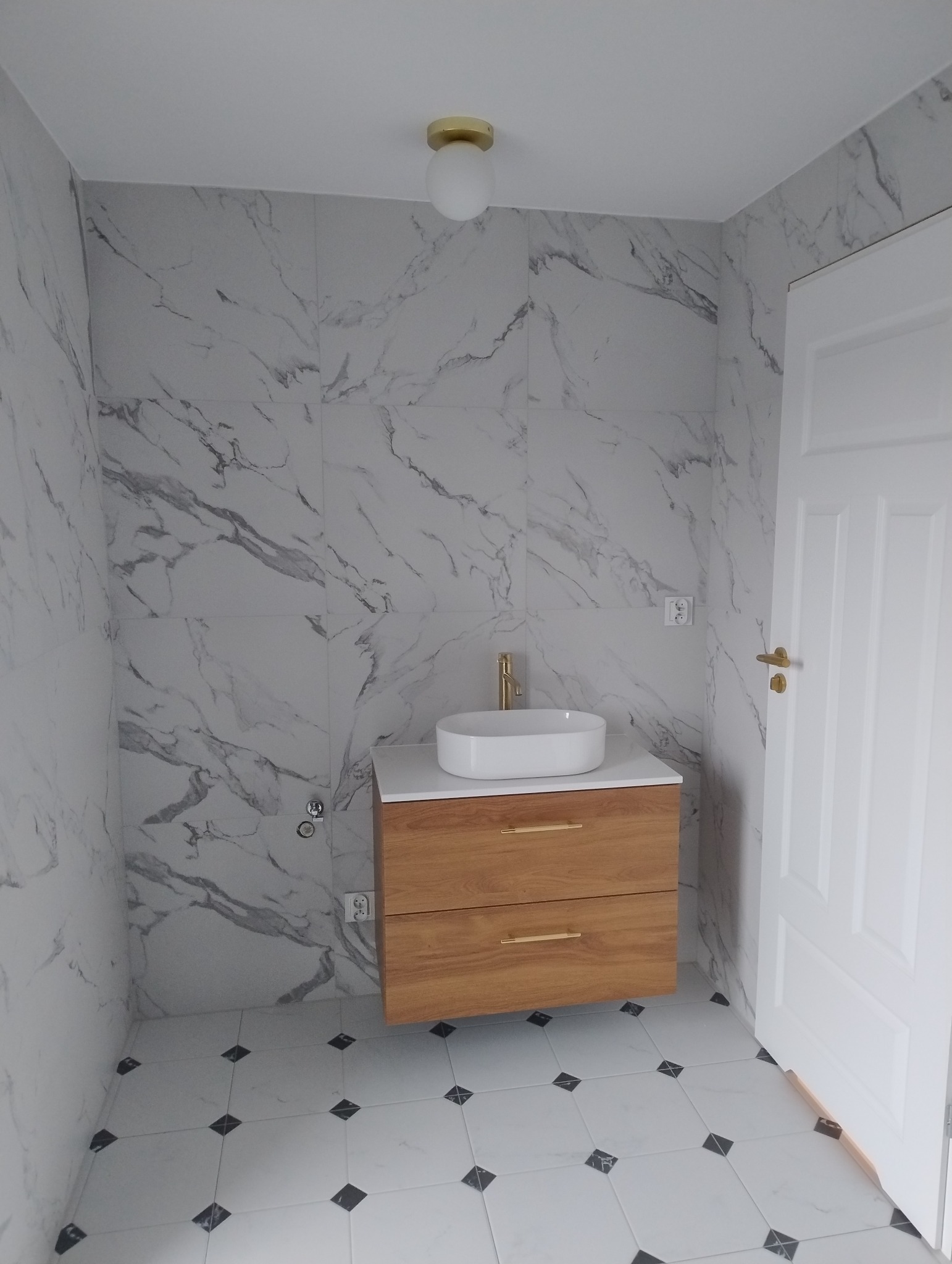 Nowoczesna łazienka z marmurowymi płytkami na ścianach, drewnianą szafką pod umywalką z złotymi uchwytami i baterią, oraz białą umywalką nablatową, oświetlona okrągłą lampą sufitową.