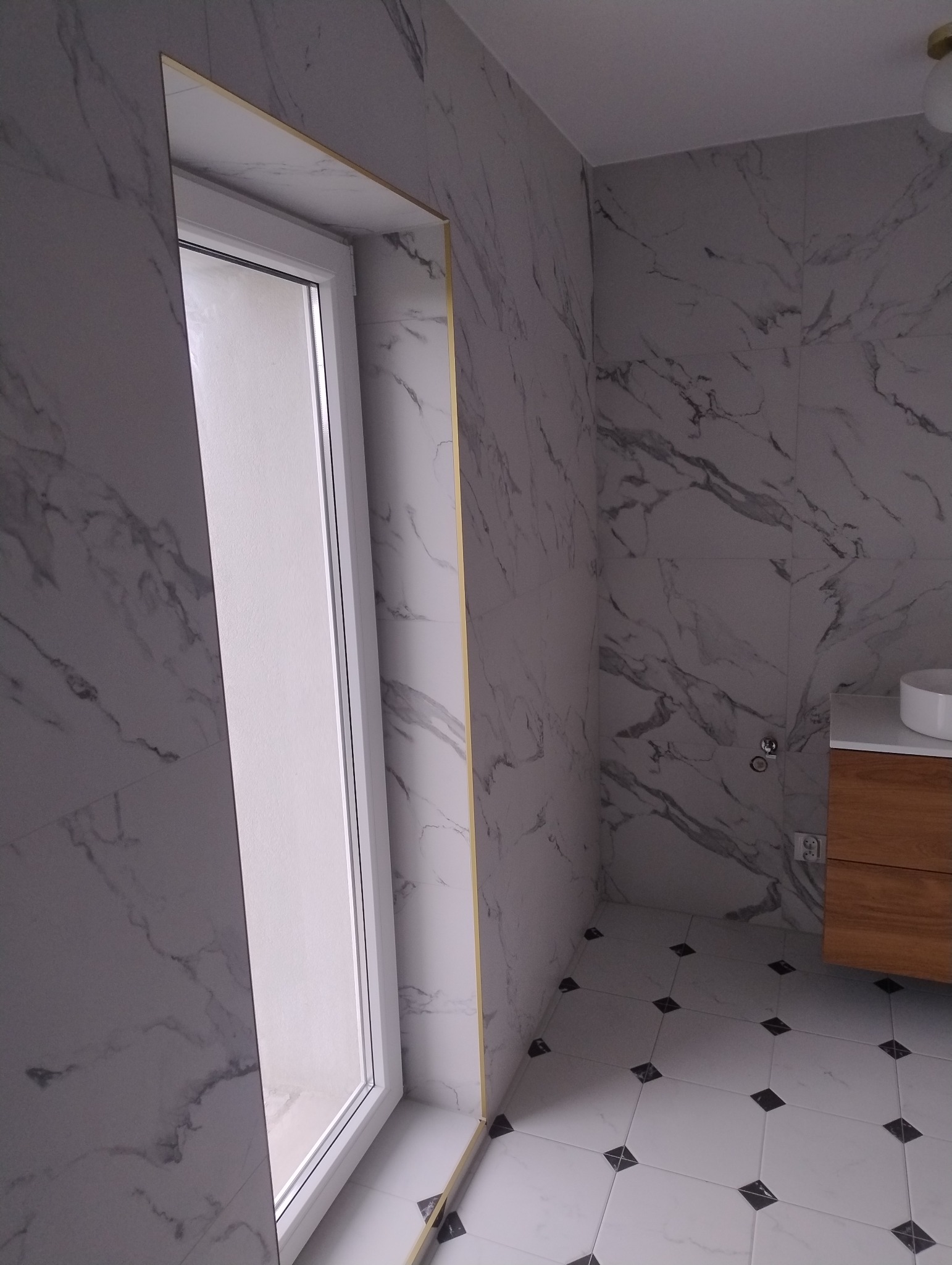 Wykończona łazienka w marmurze z białą stolarką okienną i złotymi listwami wykończeniowymi, widok na umywalkę z drewnianą szafką i biało-czarną podłogę.