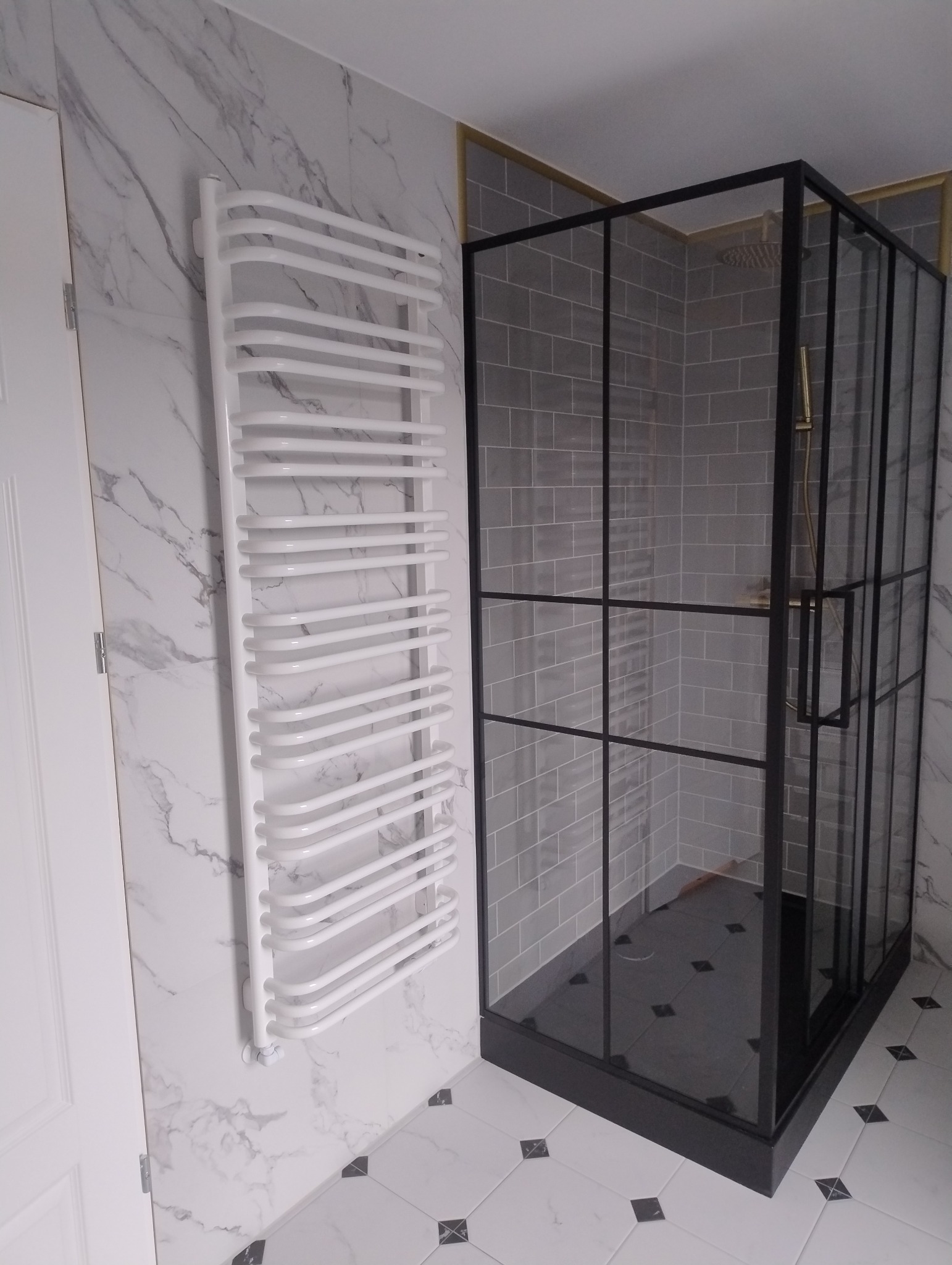Nowoczesna kabina prysznicowa z czarnymi profilami, szare płytki na ścianach i podłodze, biały grzejnik drabinkowy na ścianie z marmurowym wzorem, biała podłoga z czarnymi akcentami.