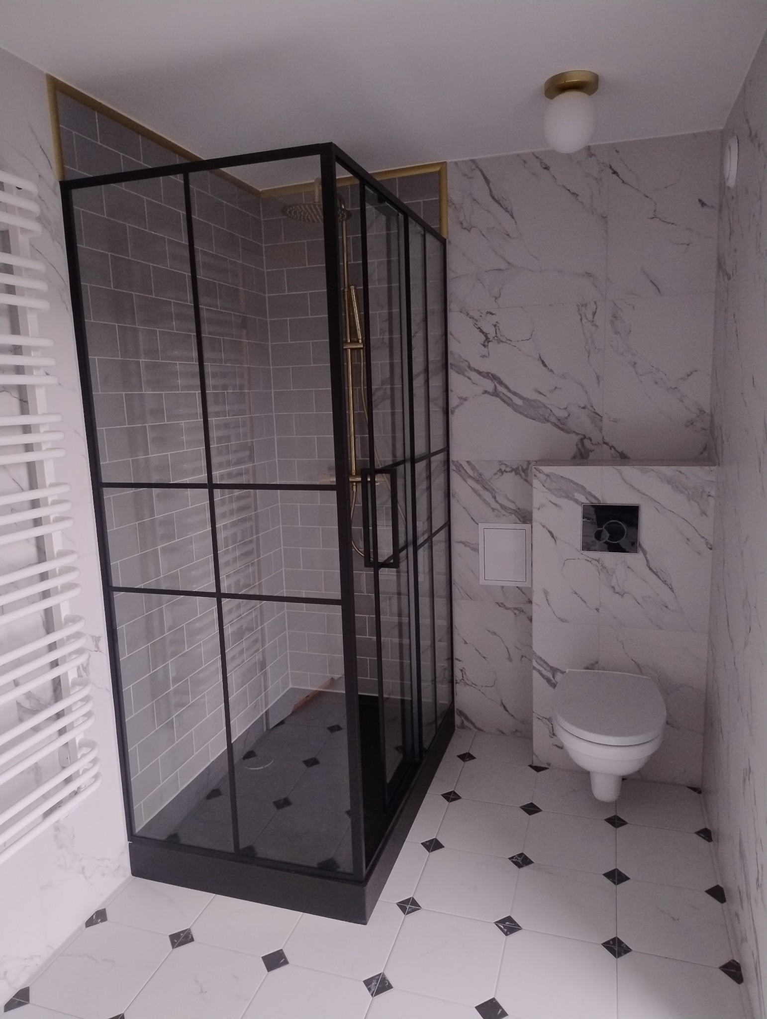 Nowoczesna łazienka z kabiną prysznicową typu walk-in z czarnymi profilami, szare płytki w kabinie, białe płytki imitujące marmur na ścianach i podłodze z czarnymi akcentami, biały grzejnik...