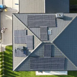 Widok z góry na dach domu z zainstalowanymi panelami słonecznymi, biały bus z otwartymi drzwiami i rampą obok, dwie osoby stojące na chodniku, trawnik i mały budynek gospodarczy w tle.