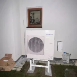 Biała zewnętrzna jednostka pompy ciepła Samsung zamontowana na betonowym fundamencie, widoczna na tle białej ściany budynku z małym oknem z brązową ramą, obok leżą kartony i opakowania.