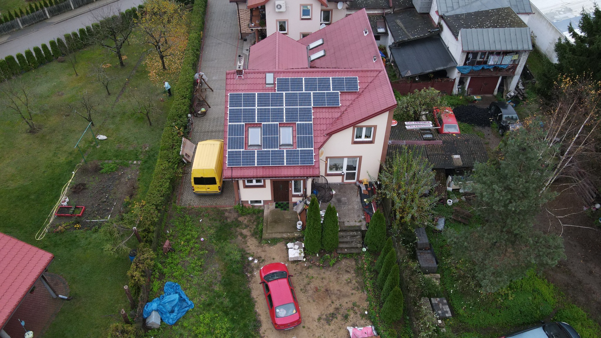 Widok z góry na dom jednorodzinny z panelami słonecznymi na dachu, żółty van zaparkowany obok, czerwony samochód na podwórku, otoczenie z zielenią i innymi budynkami.