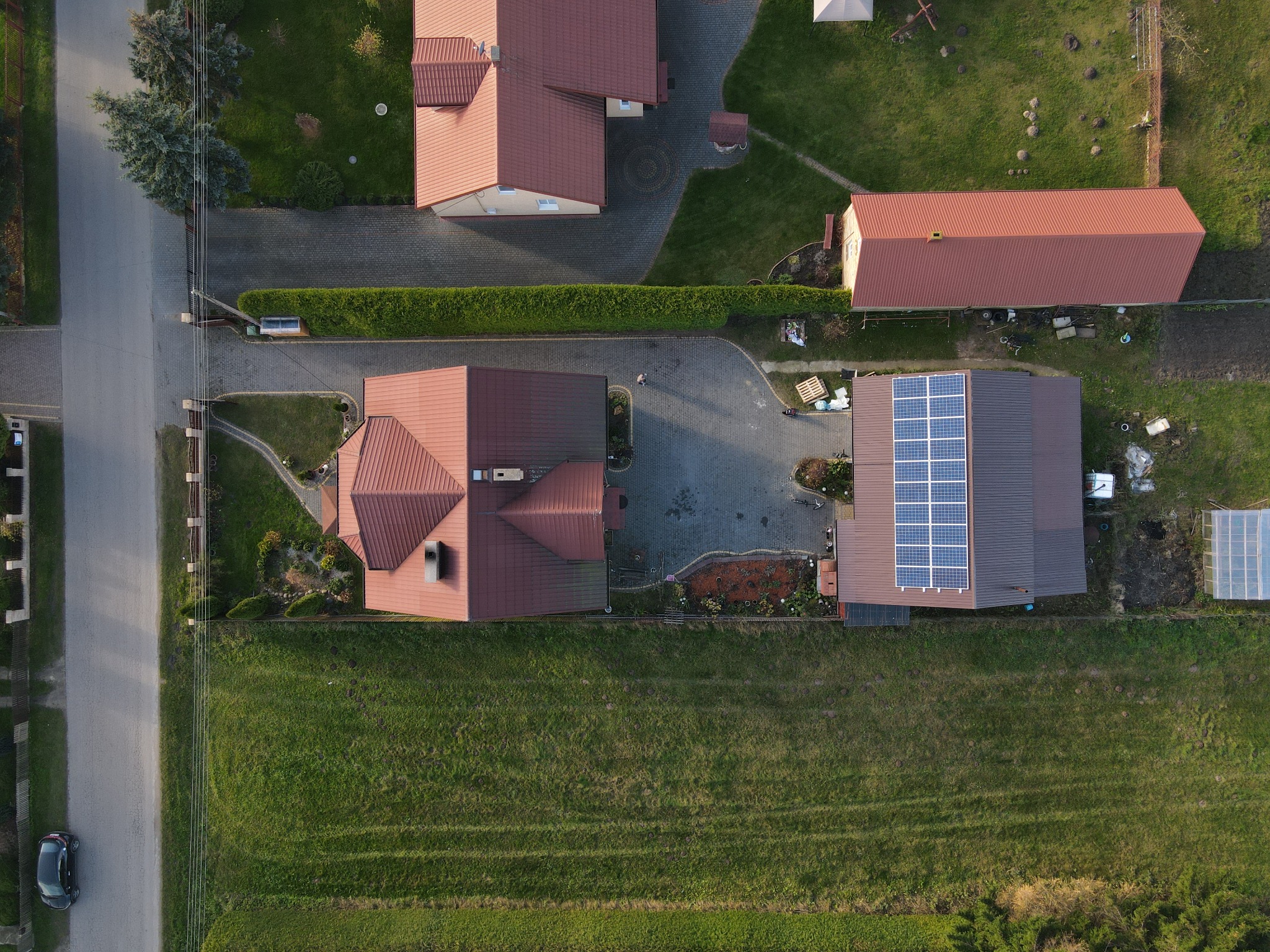 Widok z drona na domy jednorodzinne, jeden z nich z panelami słonecznymi na dachu, otoczone zielenią i drogą.